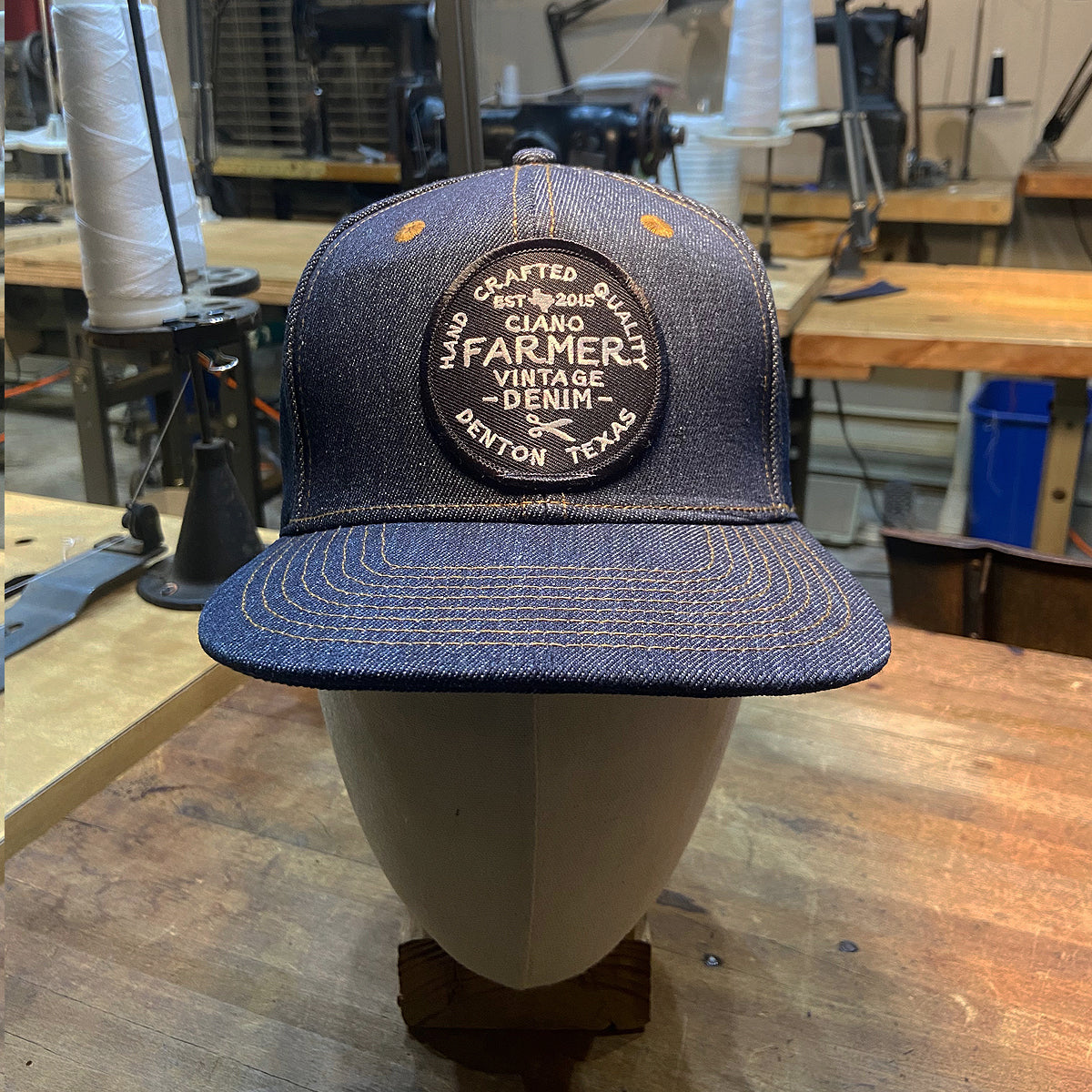 A blue denim trucker hat with 'Crafted in Texas' embroidered on the front, displayed on a mannequin head in a workshop setting.