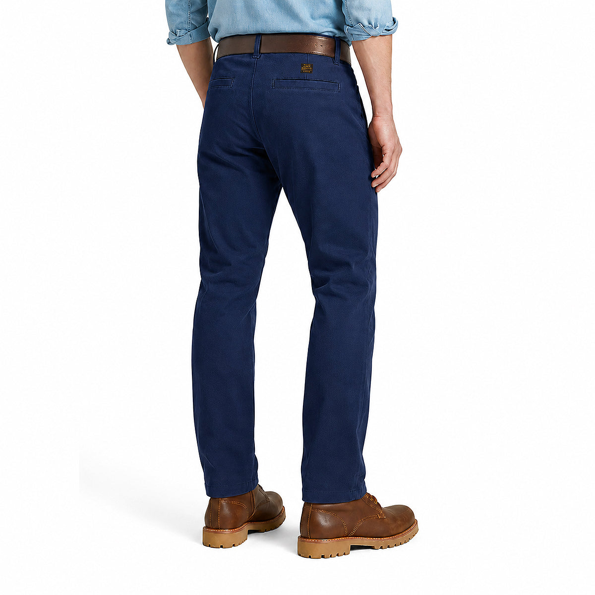 A person shown from the back wearing navy blue chino pants, a light blue button-down shirt, and brown leather boots against a clean white studio background.