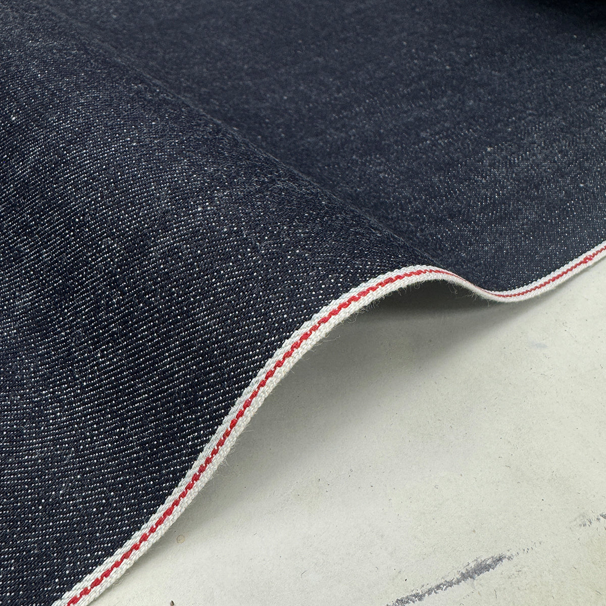 A close-up of dark indigo denim fabric with a folded edge showing a white and red contrast stitched border, placed on a light gray surface.