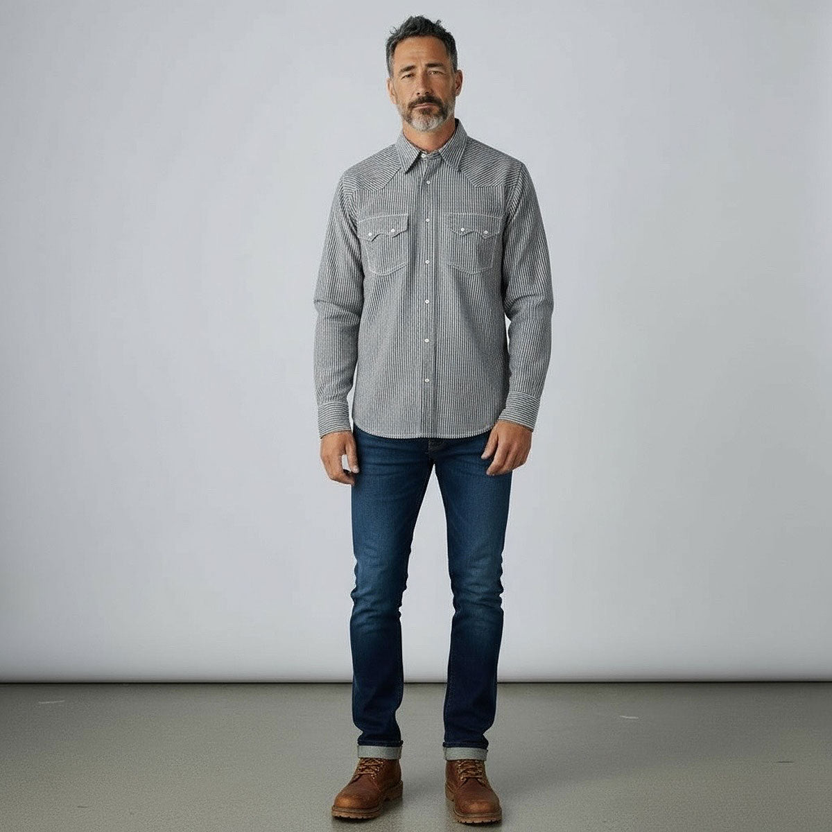 Man wearing a hickory stripe shirt, blue jeans, and brown shoes against a plain background