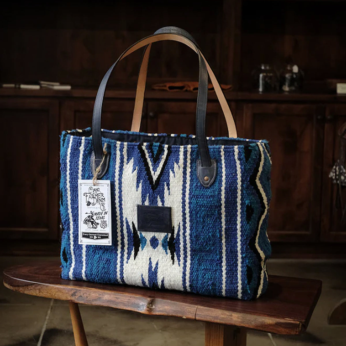 A hand woven indigo-dyed wool tote bag with leather straps, displayed on a wooden stool.