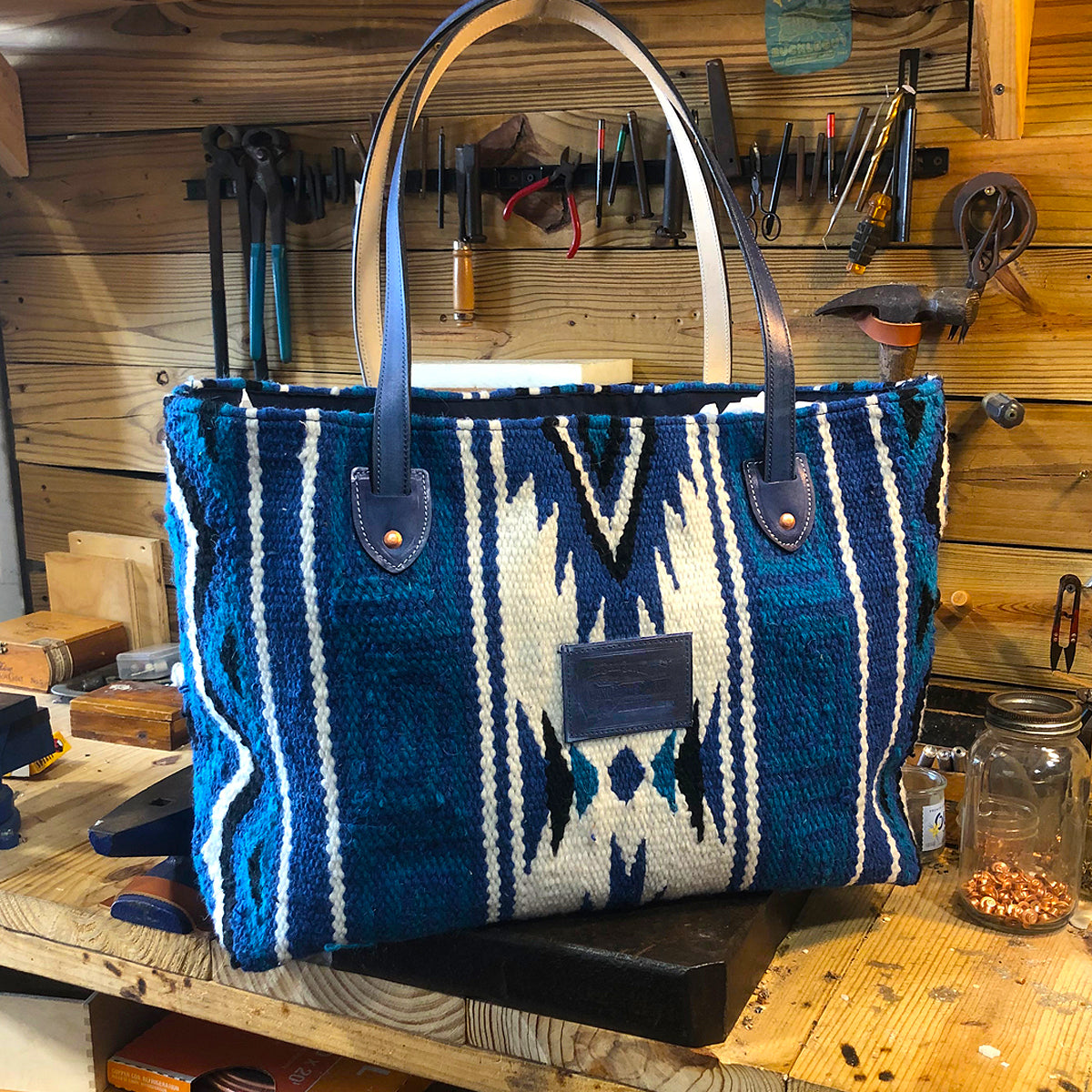 A hand woven indigo-dyed wool tote bag with leather straps, displayed on a wooden stool.